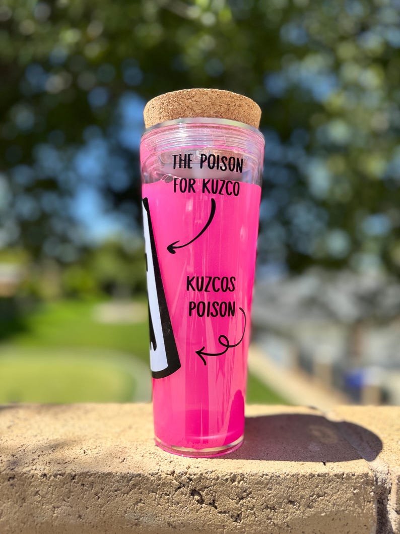 May include: A clear glass tumbler with a cork lid, filled with bright pink liquid. The text "THE POISON FOR KUZCO" is printed on the top, with "KUZCOS POISON" and an arrow on the side. The tumbler is sitting on a brick surface.