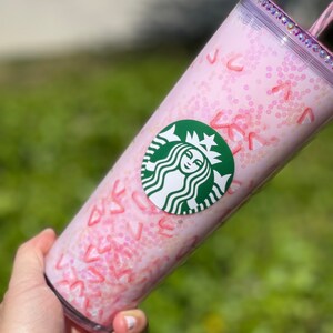 Peut inclure: Gobelet r&eacute;utilisable rose avec un logo Starbucks vert et un design de paillettes rose et blanc. Le gobelet a un couvercle transparent avec un bord de paillettes rose et blanc.