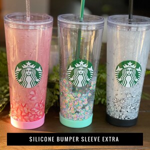 May include: Three clear plastic tumblers with silicone sleeves. The tumblers feature a Starbucks logo and are filled with glitter. The sleeves are pink, teal, and black.