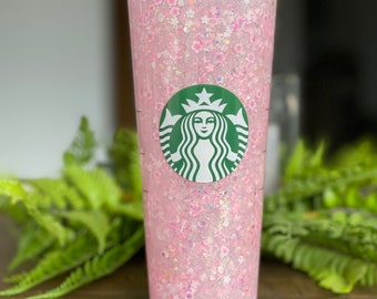 Może przedstawiać: Przezroczysty plastikowy kubek z różowo-białym wzorem z brokatem i zielonym logo Starbucks. Kubek ma słomkę.
