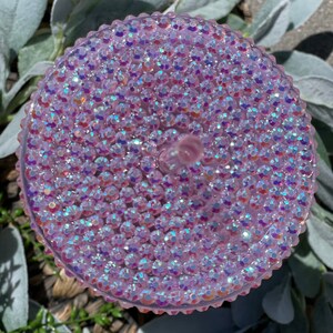 Peut inclure: Un couvercle rond et textur&eacute; de couleur rose iris&eacute;e, recouvert de petits strass scintillants.