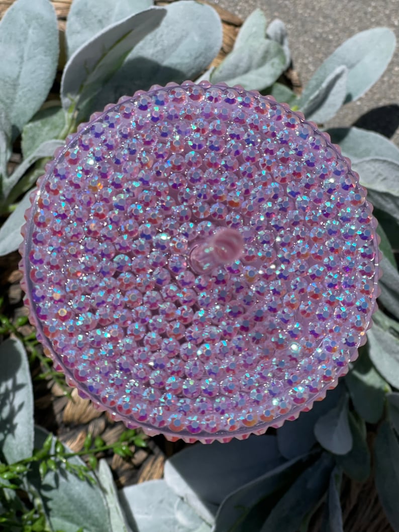 Peut inclure: Un couvercle rond et textur&eacute; de couleur rose iris&eacute;e, recouvert de petits strass scintillants.