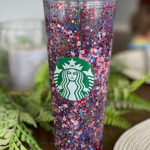 May include: A clear plastic tumbler with a green Starbucks logo and red, white and blue glitter inside. The tumbler has a lid and a straw.