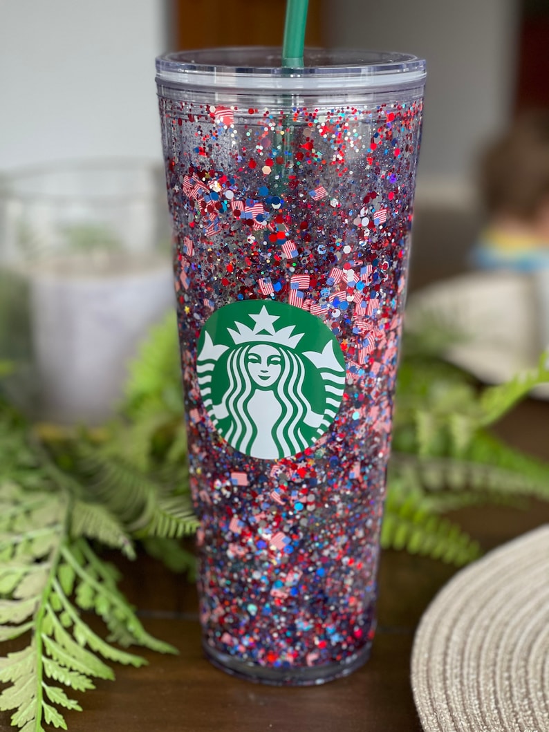 May include: A clear plastic tumbler with a green Starbucks logo and red, white and blue glitter inside. The tumbler has a lid and a straw.