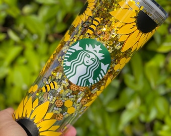 May include: A clear tumbler with a gold glitter background and a design of sunflowers, bees, and honeycombs. The tumbler also features a green Starbucks logo.