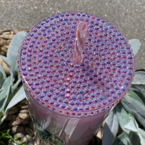 Peut inclure: Un gobelet en plastique transparent avec un couvercle rose iridescent recouvert de strass. Une paille rose est ins&eacute;r&eacute;e dans le couvercle.