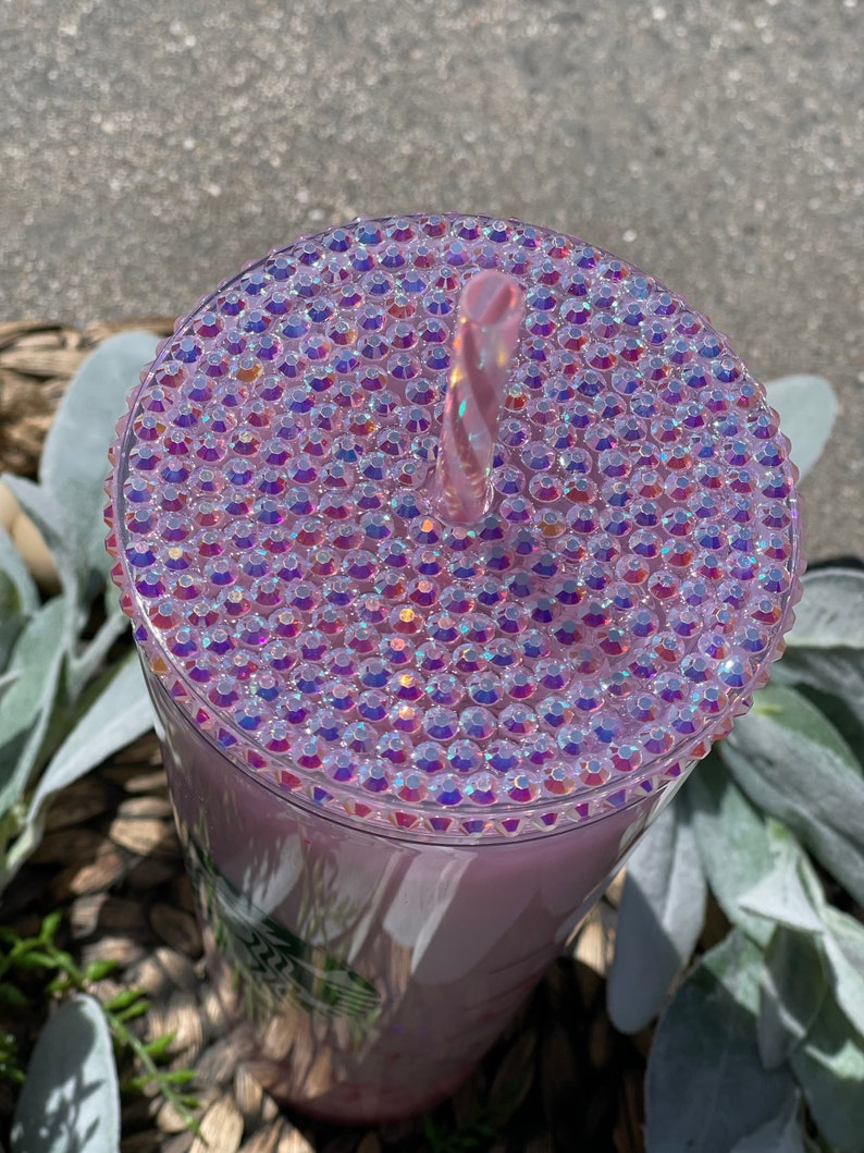 Peut inclure: Un gobelet en plastique transparent avec un couvercle rose iridescent recouvert de strass. Une paille rose est ins&eacute;r&eacute;e dans le couvercle.
