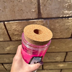 May include: A clear, pink tumbler with a cork lid. The tumbler has a black and white label with the letter "M" visible. The cork lid has a circular hole in the center. The tumbler is held in front of a brick wall.