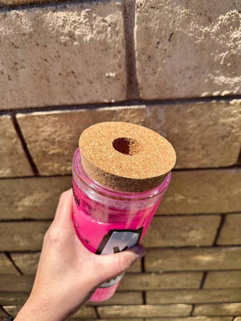 May include: A clear, pink tumbler with a cork lid. The tumbler has a black and white label with the letter "M" visible. The cork lid has a circular hole in the center. The tumbler is held in front of a brick wall.