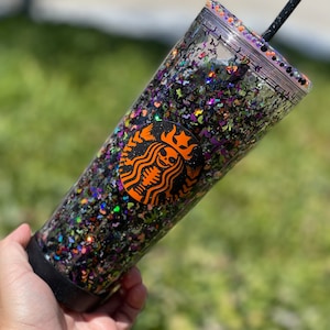 May include: A clear plastic tumbler with a black lid and a black straw. The tumbler is filled with black, orange, green, and purple glitter. A black and orange Starbucks logo with a skeleton inside is visible on the side of the tumbler.