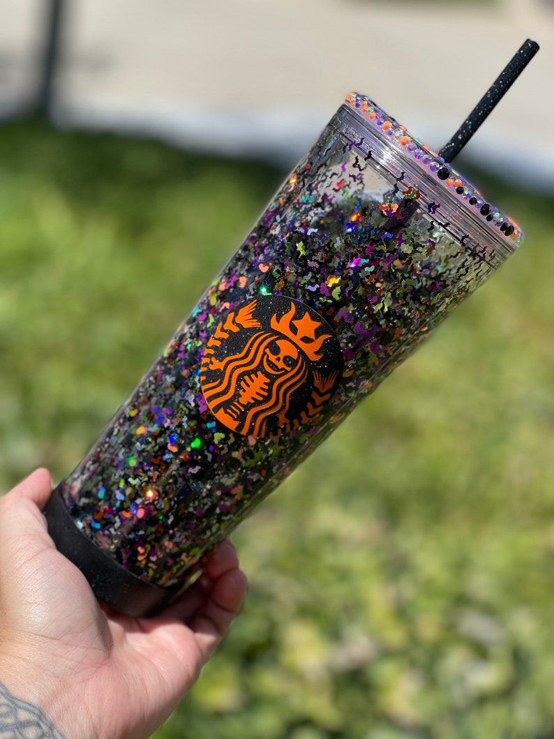 May include: A clear plastic tumbler with a black lid and a black straw. The tumbler is filled with black, orange, green, and purple glitter. A black and orange Starbucks logo with a skeleton inside is visible on the side of the tumbler.