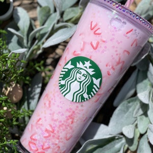 Peut inclure: Un gobelet r&eacute;utilisable rose avec un logo Starbucks vert et un design de paillettes rose et blanc. Le gobelet a un couvercle transparent et une paille.