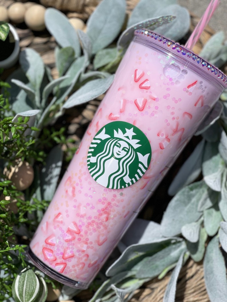 Peut inclure: Un gobelet r&eacute;utilisable rose avec un logo Starbucks vert et un design de paillettes rose et blanc. Le gobelet a un couvercle transparent et une paille.