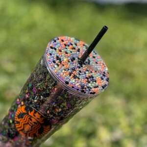 May include: A clear reusable tumbler with a black glitter and orange, green, and purple iridescent glitter design. The lid is decorated with black, orange, and green rhinestones. A black straw is inserted into the lid.