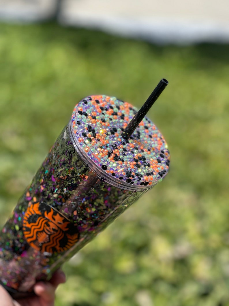 May include: A clear reusable tumbler with a black glitter and orange, green, and purple iridescent glitter design. The lid is decorated with black, orange, and green rhinestones. A black straw is inserted into the lid.