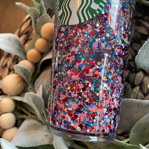May include: A clear tumbler with a green Starbucks logo on the top. The tumbler is filled with red, white and blue glitter and small American flag shapes.