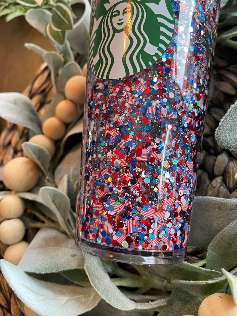 May include: A clear tumbler with a green Starbucks logo on the top. The tumbler is filled with red, white and blue glitter and small American flag shapes.