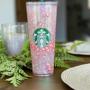 May include: A pink and clear reusable tumbler with a green straw and a Starbucks logo. The tumbler is filled with pink glitter and small pink and white strawberry shapes.