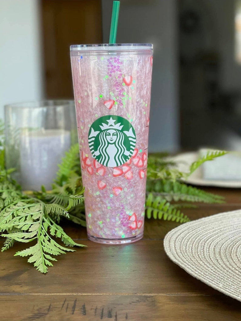 May include: A pink and clear reusable tumbler with a green straw and a Starbucks logo. The tumbler is filled with pink glitter and small pink and white strawberry shapes.