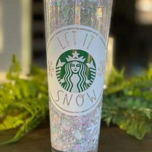 May include: A clear tumbler with a white lid and a white snowflake design. The tumbler is filled with white glitter and has a green Starbucks logo with the text "Let it Snow" around it.
