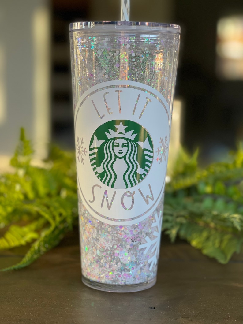 May include: A clear tumbler with a white lid and a white snowflake design. The tumbler is filled with white glitter and has a green Starbucks logo with the text "Let it Snow" around it.