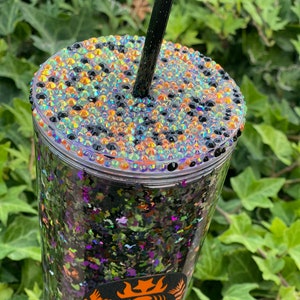 May include: A clear plastic tumbler with a black and green glitter design. The tumbler has a black straw and a lid with a black, orange, and iridescent rhinestone design. The tumbler has a Starbucks logo with a skeleton design.