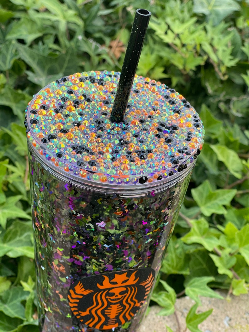 May include: A clear plastic tumbler with a black and green glitter design. The tumbler has a black straw and a lid with a black, orange, and iridescent rhinestone design. The tumbler has a Starbucks logo with a skeleton design.