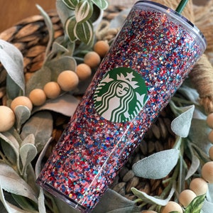 May include: A clear tumbler with a red, white and blue glitter design. The tumbler has a green Starbucks logo on the front.