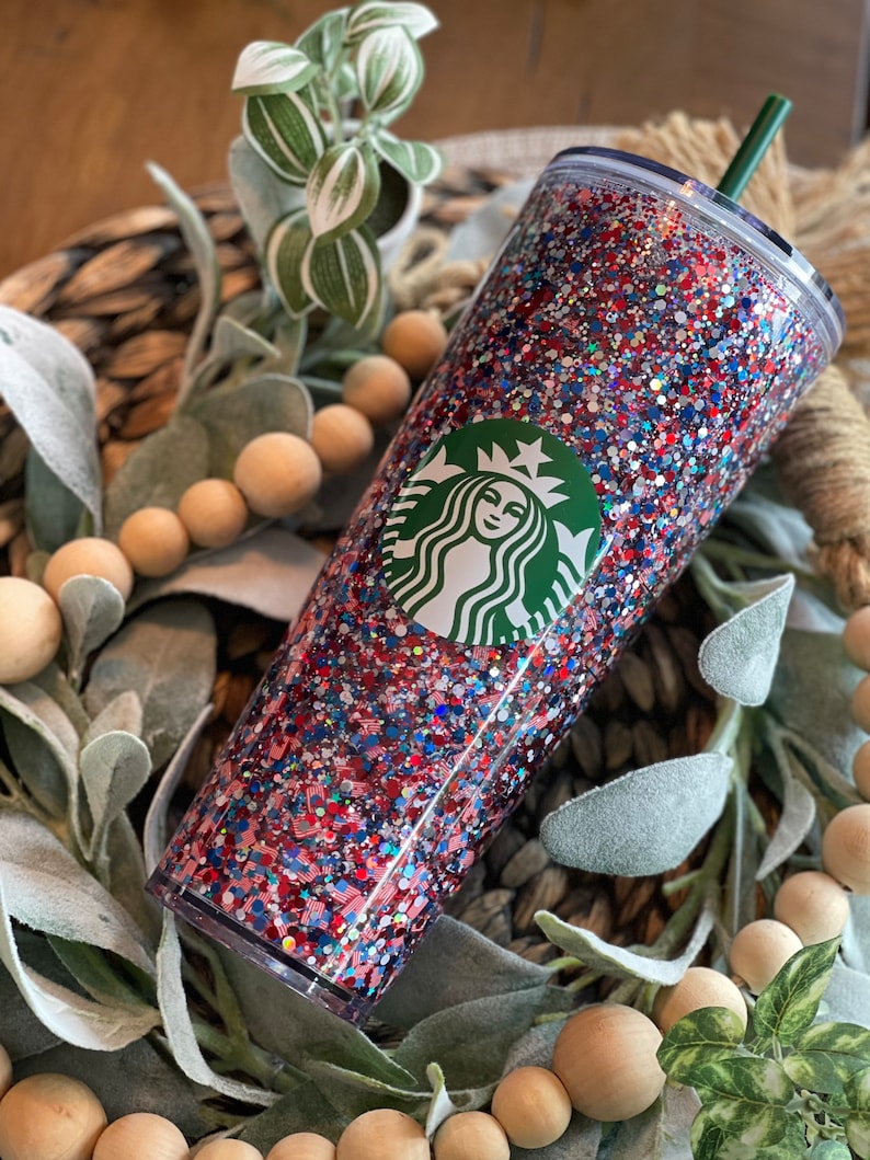 May include: A clear tumbler with a red, white and blue glitter design. The tumbler has a green Starbucks logo on the front.
