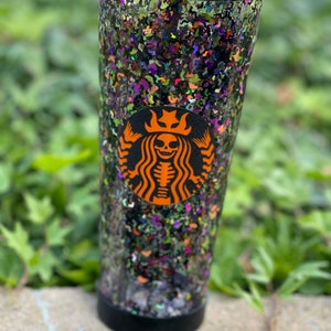 May include: A clear tumbler with a black lid and a black Starbucks logo featuring a skeleton with a crown. The tumbler is filled with black, green, purple, and orange glitter.