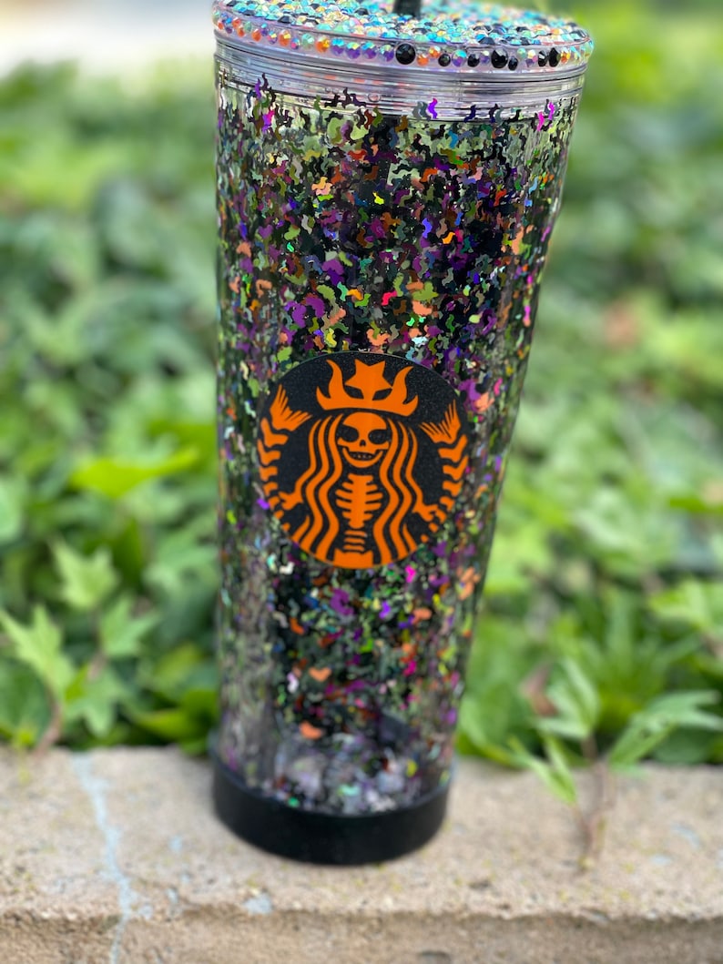 May include: A clear tumbler with a black lid and a black Starbucks logo featuring a skeleton with a crown. The tumbler is filled with black, green, purple, and orange glitter.