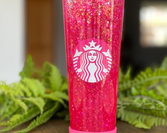 May include: A pink tumbler with a clear lid and a pink silicone base. The tumbler is filled with pink glitter and has a white Starbucks logo on the side.