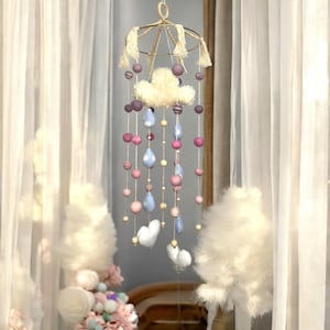 May include: A handcrafted baby mobile featuring a gold-colored metal frame with hanging strings of pink, purple, and blue felt balls, teardrops, and cloud shapes. The mobile is suspended in front of a sheer white curtain.