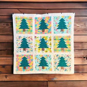 May include: A handmade quilt with a repeating pattern of teal Christmas trees on a white background. Each tree is set against a colorful square of floral and plaid patterns. The quilt is hanging on a wooden wall.