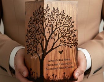 May include: A wooden urn with a tree design, butterflies, and the text "Always In Our Hearts." The urn is engraved with the name Richard F. Mitchell, and the years 1943-2026. The urn is held by a person.