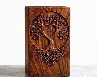 May include: A rectangular wooden cremation urn featuring a deep brown grain and a circular relief carving on the front. The carving depicts a tree with intricate roots and branches forming a heart shape in the center with the word Family engraved.