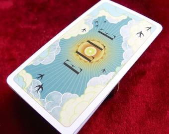 Op de afbeelding: Een pak tarotkaarten met een hemels ontwerp. De achterkanten van de kaarten hebben een zonnestraalontwerp met wolken, vogels en het woord "L E" in zwart. De kaarten hebben witte randen en liggen op een rode fluwelen ondergrond.