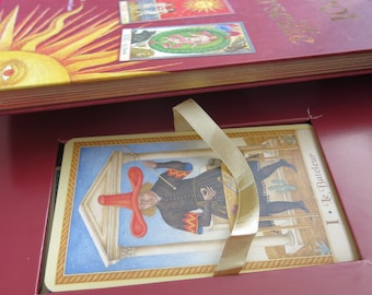 May include: A red box with a gold ribbon and a tarot card inside. The card is titled "I Le Bateleur" and features a man in a red hat and a blue and gold costume standing in front of a building.