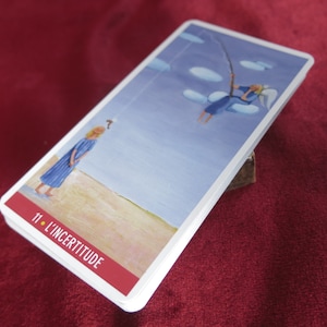 Op de afbeelding: Een tarotkaart met een blauwe achtergrond en een vrouw in een blauwe jurk die op een strand staat. De vrouw kijkt omhoog naar een figuur in de lucht, die met een hengel vist. De kaart is getiteld "11. L'Incertitude".