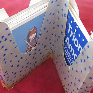 Könnte beinhalten: Ein weißer Kartenhalter mit blauen Sternmustern, der ein Kartenspiel enthält. Die Karten haben Illustrationen und Text. Eine Seite des Halters trägt den Text "Trion Amore". Die andere Seite hat eine Illustration einer Person.