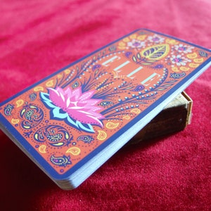 May include: A deck of cards with a colourful paisley design and the word "ELLE" printed on the front. The card features a pink lotus flower with blue and green leaves.