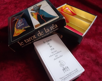 Puede incluir: Una caja negra de cartas del tarot con interior dorado y rojo, y una baraja de cartas. La tapa de la caja presenta ilustraciones y el texto "le tarot de la réa". Las cartas son blancas con texto negro y un logotipo en espiral.
