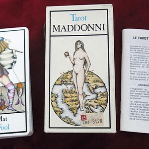 May include: Tarot deck titled "Maddonni" with a card depicting "The Fool". The box shows a nude woman holding an apple on a world map. A booklet with French text is also included.