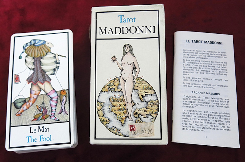 May include: Tarot deck titled "Maddonni" with a card depicting "The Fool". The box shows a nude woman holding an apple on a world map. A booklet with French text is also included.