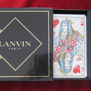 Puede incluir: Una caja negra con un diseño dorado que presenta el texto "LANVIN PARIS" y una imagen estilizada de un bufón. La caja contiene dos cartas de juego, la Reina de Corazones, con un diseño de lunares rojos y blancos, y la Reina de Espadas, con un diseño de lunares azules y blancos.