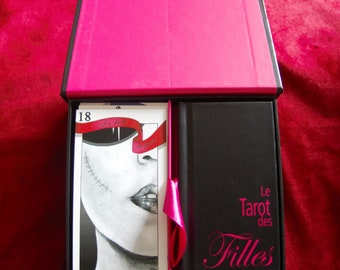 May include: A black box with a pink lid and a pink ribbon. The box contains a deck of tarot cards with a black and white illustration of a woman's face on the card. The box also has the text "Le Tarot des Filles" on the front.