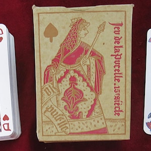 Puede incluir: Una baraja de cartas vintage con ilustraciones de figuras históricas. La caja de cartas y algunas cartas presentan texto en francés, incluyendo "Jeu de la Pucelle 15 siècle". Las cartas tienen un diseño clásico.