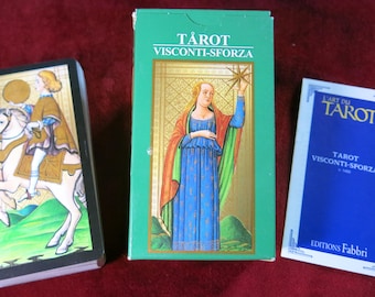 May include: A green box with the text "TÅROT VISCONTI-SFORZA" on the front. The box contains a deck of tarot cards featuring illustrations of people and scenes from the Visconti-Sforza tarot deck. The box is sitting on a red surface.