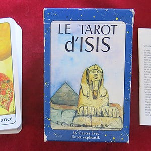 Könnte beinhalten: Ein Tarotkartenset mit einer blauen Schachtel mit der Aufschrift "LE TAROT d'ISIS" und einer Abbildung der Sphinx und einer Pyramide. Ein Stapel Karten mit einer Horn- und Herzillustration und dem Text "Corne d'abondance" befindet sich neben der Schachtel. Ein weißes Büchlein ist ebenfalls vorhanden.