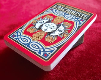 Puede incluir: Una baraja de cartas del tarot con borde rojo y diseño trasero. La parte trasera presenta tres figuras con caras de sol con detalles dorados y azules, y el texto "GRAND TAROT DIVINATOIRE L'ALCHIMISTE". La baraja descansa sobre una superficie marrón oscuro.
